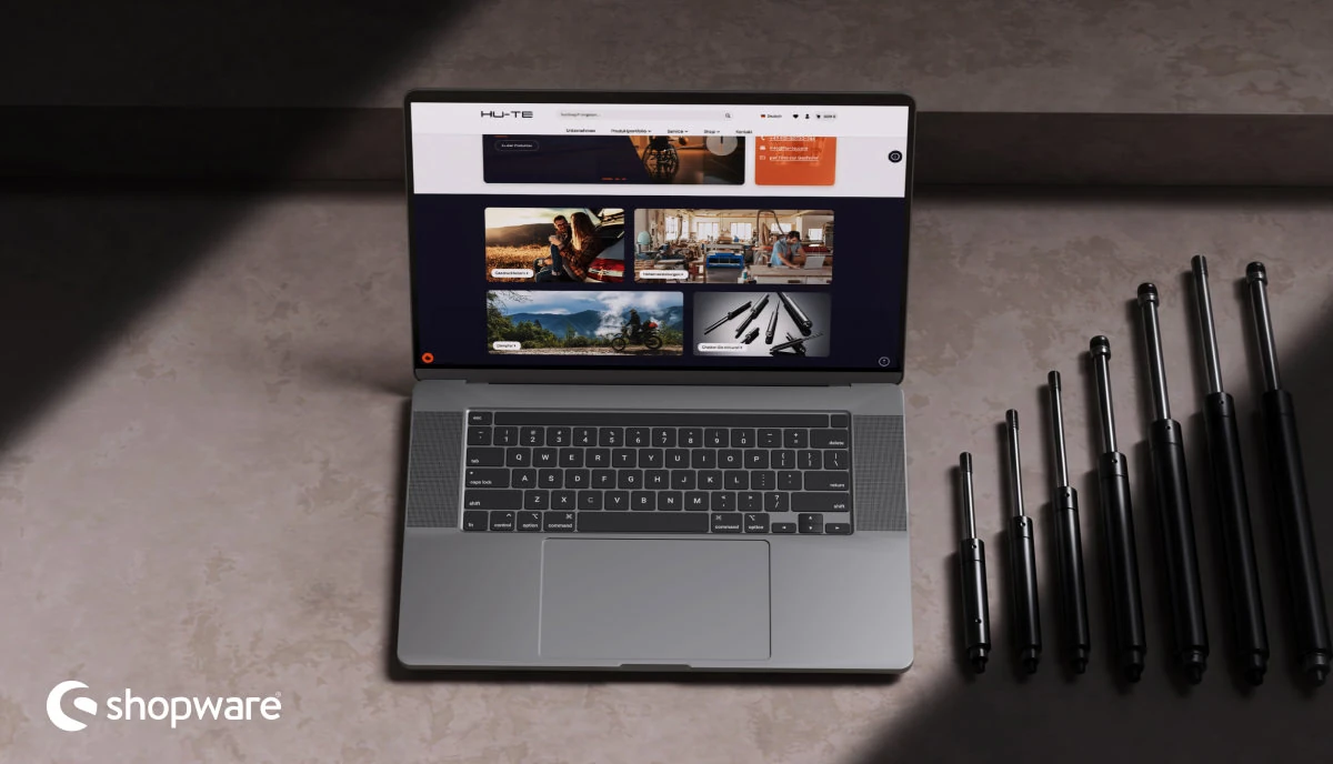 Shopware-6-Onlineshop auf einem Laptop neben mehreren schwarzen Gasdruckfedern auf dunkler Arbeitsfläche.