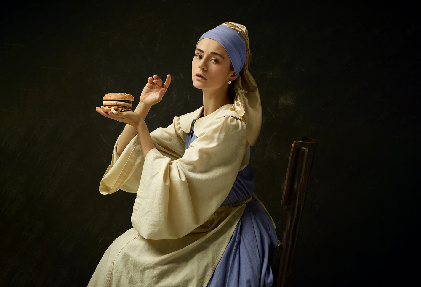 Frau, die wie „Das Mädchen mit dem Perlenohrring“ von Jan Vermeer gestyled ist, hält kunstvoll einen Hamburger in der Hand.