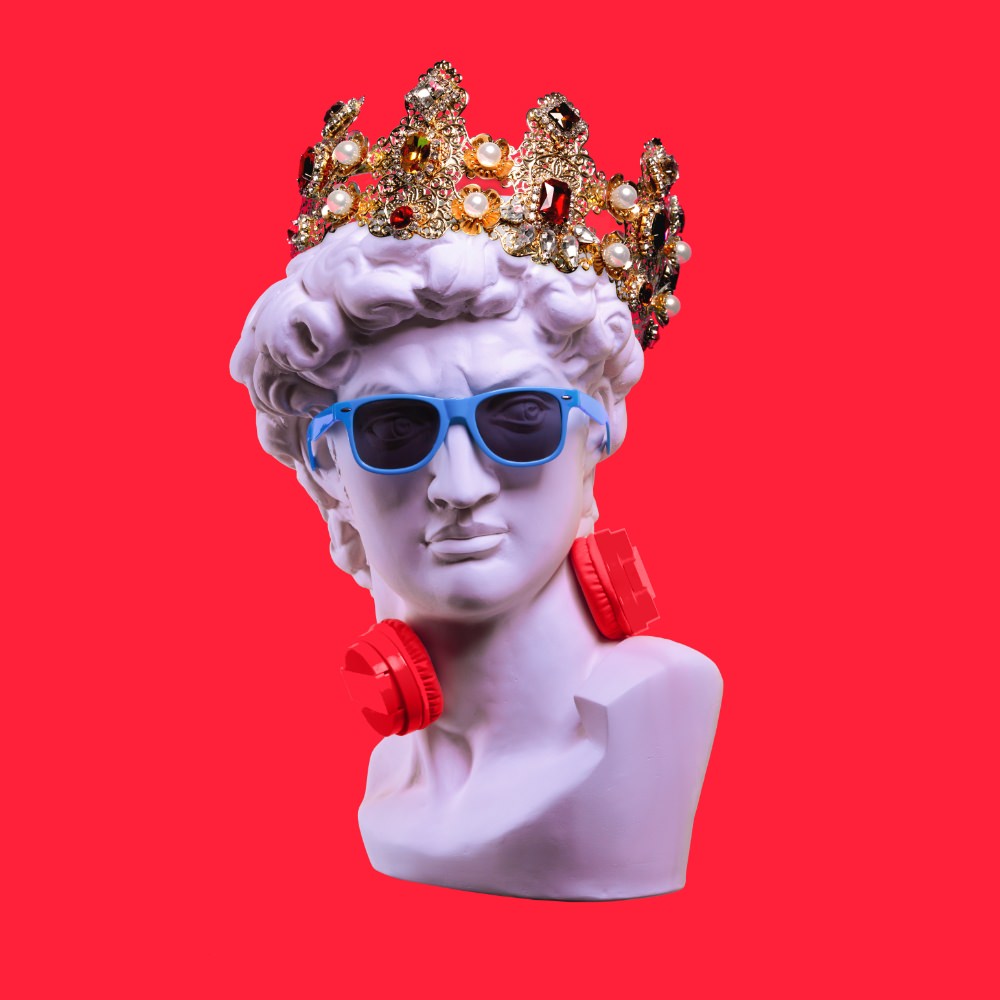 Büste einer antiken Statue mit goldener Krone, blauer Sonnenbrille und roten On-Ear-Kopfhörern um den Hals.